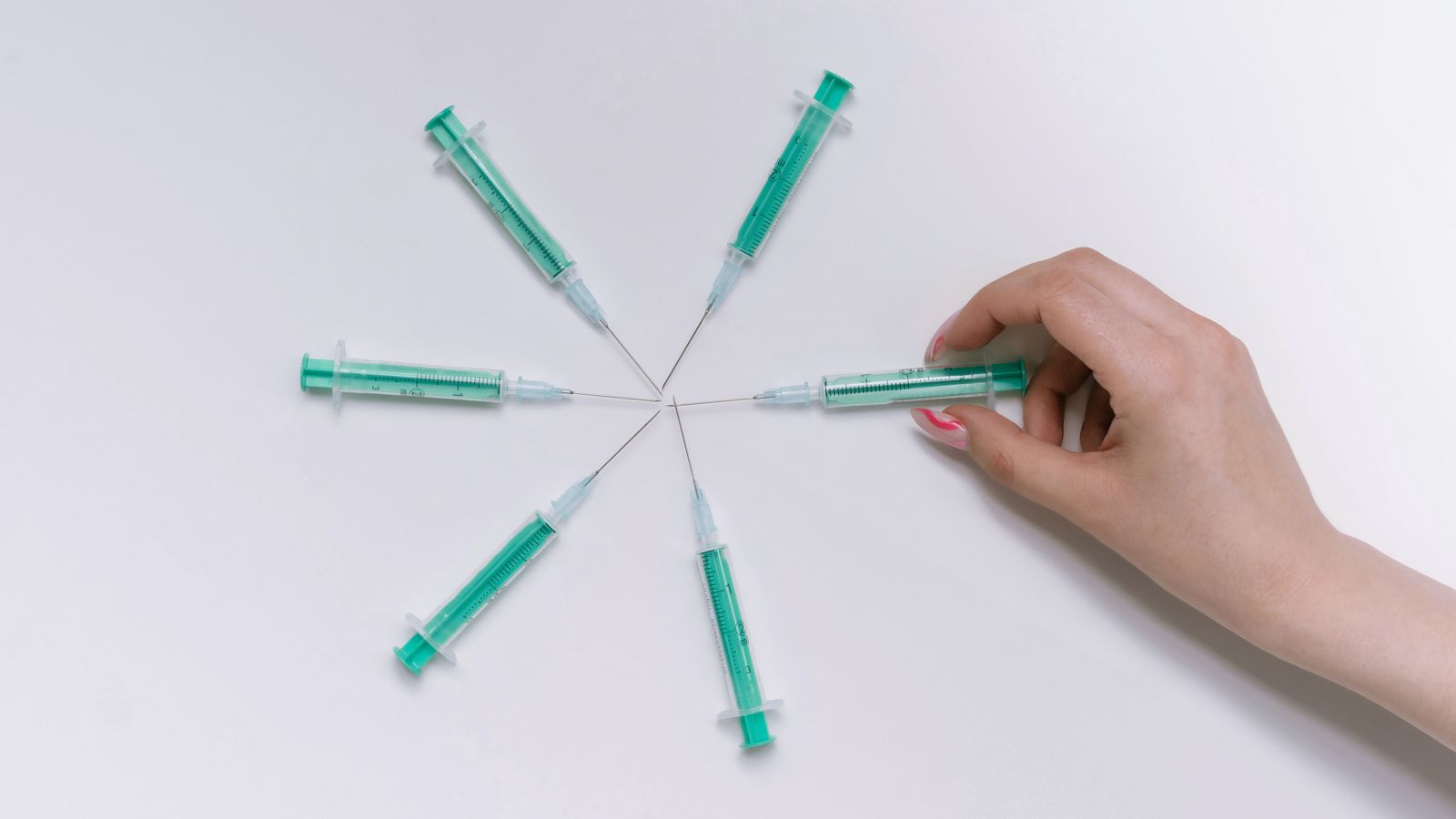 A hand arranges green syringes in a circular pattern on a white surface.