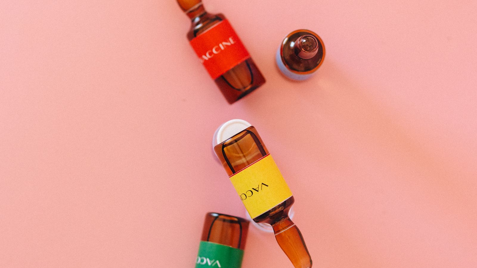 Four small vials with colored labels arranged on a pastel pink background.