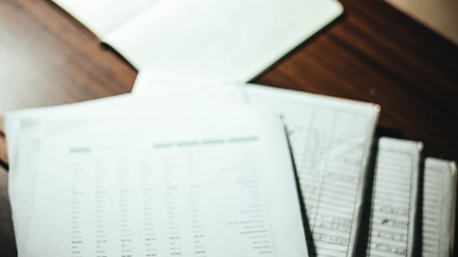 An overhead view of a stack of documents covered in statistics and data, resting on a desk beside an open book and a cup of a hot drink in a dimly lit room.